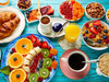 Reichhaltiges Frühstücksbuffet mit frischen Früchten, Croissants, Kaffee, Orangensaft, Müsli und Trockenfrüchten auf blauem Holztisch.